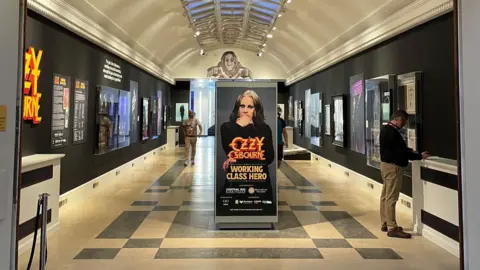 An art gallery hall with framed pictures on black walls. There is a poster of a man in the centre of the room, which says "Ozzy Osbourne, Working Class Hero" on it.