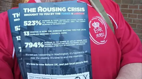 Wearside Reform UK Close up shot of blue leaflet being held by a man wearing a red T-shirt which says on the breast pocket "British Army Rugby" with an army logo. The leaflet shows various statistics including "794% increase in the number of asylum seekers being housed in Sunderland, with the the biggest increases occurring after 'City of Sanctuary' introduced".