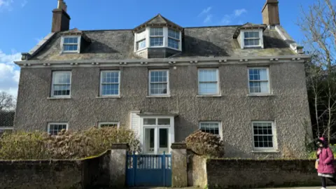 BBC A large rectory house in St Martin, Guernsey. It is a detached grey-coloured building with several sash windows, a stone fence and a blue gate. A woman wearing a purple coat is walking on the pavement.