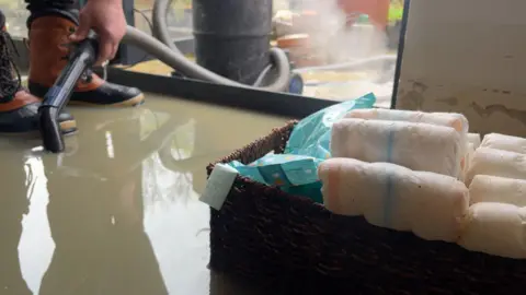 Harry Parkhill/BBC A box of nappies can be seen amid at least two inches of brown floodwater. A person wearing boots and holding a hose is pumping water from the house.