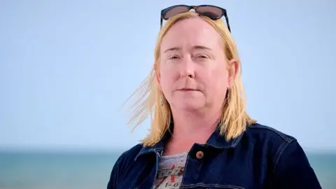 A woman with blond shoulder-length hair, standing on a pebble beach with the sea in the background. She is wearing a blue denim jacket. The sky is blue and cloudless.