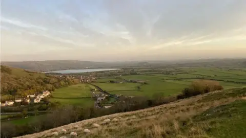 A panorama of a distant Cheddar reservoir surrounded by green fields with a hillside of sheep in the foreground. Several houses are dotted in the landscape.