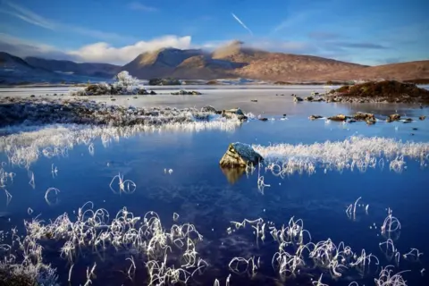 Peter Xu A loch that has frozen over and is now ice, with hills in the distance and a clear blue sky above