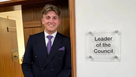 George Finch leans against a wall. He is wearing a navy jacket and purple toe and white shirt. A sign to the right of the image reads "Leader of the Council"