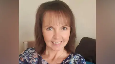 Heather Smedley, who has brown shoulder length hair, smiles while sat on a sofa.