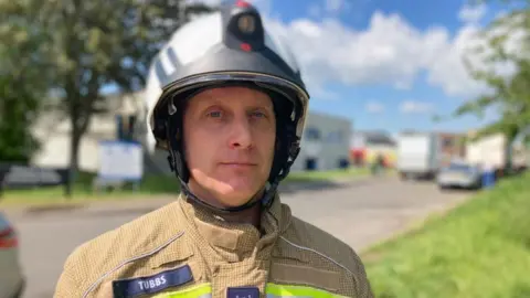 Alex Pope/BBC A firefighter, wearing a brown uniform, with a helmet, standing by a building, with grass banks behind him. A tree and several buildings are behind him. 
