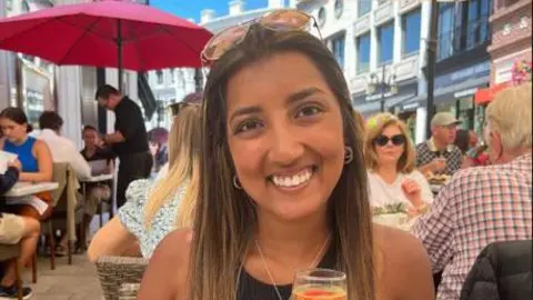 A woman with shoulder length dark hair has sunglasses on her head, and is holding a glass. She is smiling, and sat at a table, with other people sat at tables around her
