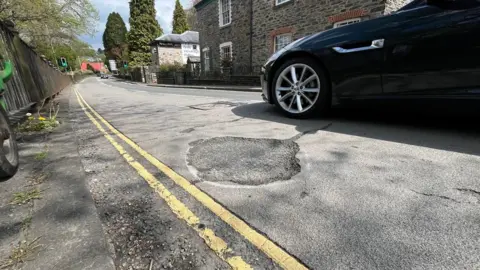 A pothole on a road next to double yellow lines and a navy blue car with chrome details drving past
