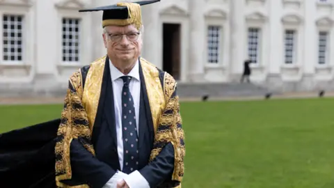 Chris Smith is wearing a black mortar board with gold trims and an ornate black and gold robe over a navy suit, white shirt and navy tie with emblems. He is standing up, with a lawn and grand building in the background.