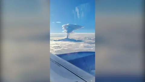 A large column of smoke rises from Mount Etna, seen from the window of a plane