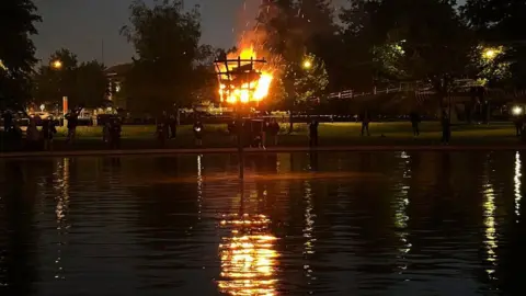 Newbury Town Council A flaming beacon reflected in the water in darkness.