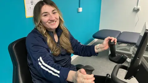 Brooke is on an arm strength piece of gym equipment against a bright blue turquoise wall. She is wearing a navy long sleeved training top and has long blond curly hair. She is smiling at the camera. 