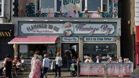 Google An ice cream shop decorated with flamingos, lots of people are seated outside.