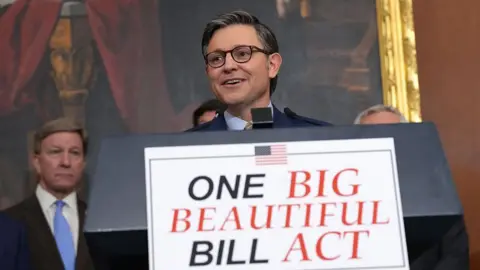 House Speaker Mike Johnson stands before a podium reading "One big beautiful bill act" for a press conference after a major House bill was passed. He is smiling.