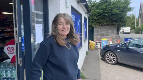 Julie Barnett stands outside a shop. She is wearing a blue jumper with a golden necklace.