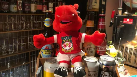 A Wrexham dragon toy perched on top of beer cans . Glasses can be seen in the background with various bottles of alcohol