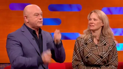 A bald man, Ross Kemp plays air drums while sitting next to Pamela Anderson, who has long blonde hair