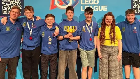 Shaun Mitchell Five boys in blue hoodies all with their arms round each other, some of them have medals. Their teacher is stood on the end and the boy in the middle is holding a yellow trophy made out of Lego.