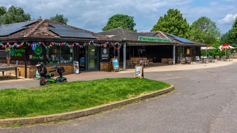 Nene Park Trust The Ferry Meadows visitor centre. A one-storey building with solar panels on the roof. There is bunting decoration above the entrance and in the distance a number of tables and chairs. There is a mobility scooter parked outside the entrance. 