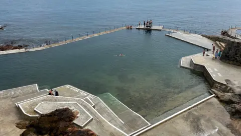 Matt Judge/BBC La Valette is an outdoor pool with a seawall enclosing some of the sea water. Steps lead down to the pool and there is some seaweed in the water. The coastline is just beyond.