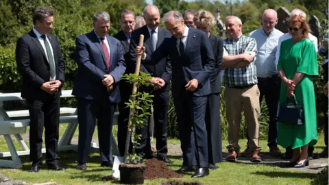 Reuters Canadian Prime Minister Mark Carney plants an 'Irish Oak' during a visit to his ancestral village Aghagower, He is holding a shovel and wearing a navy suit. There is a crowd of people behind him. 