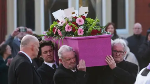 A pink glittery coffin being carried into church, with flowers and cards on top of it