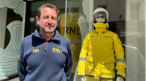 RNLI crew member, Mike Edward stands in front of the shop housing the exhibition, with a mannequin wearing RNLI uniform in the shop window behind him