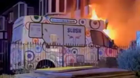 Ice cream van engulfed by flames on a driveway.