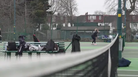 In the background, a woman with tennis racket at the net on a green court with bags and kit on benches in the foreground