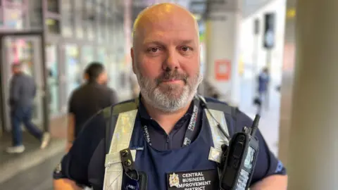 A man with a bald head and white facial hair is wearing a navy T-shirt and a navy vest with a black radio and silver reflective strips. A fabric badge on his chest reads "central business improvement district".