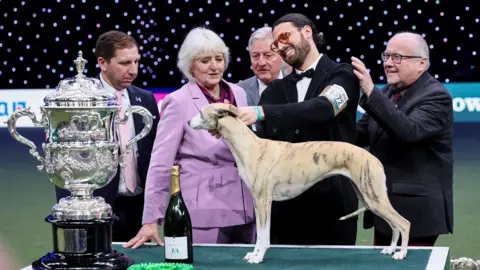 Reuters Miuccia stands on a table next to a bottle of champagne, trophy and five people celebrating