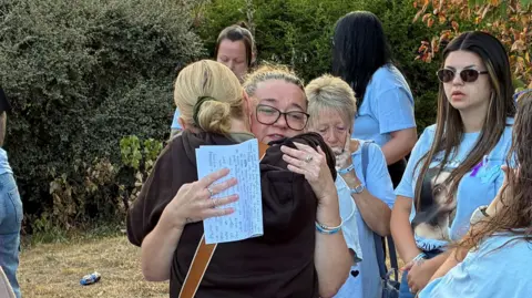 George King/BBC Two women embrace in a passionate hug as other women wearing blue T-shirts look on.