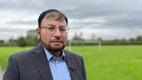 Gabby Colenso/BBC Dr Shakoor looks solemn as he looks directly at the camera. He is standing in a field, wearing a jacket and blue shirt with a microphone attached to his left lapel. He is wearing glasses.