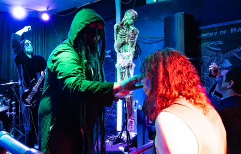 Luke Orchard Luke Orchard is on stage wearing a long black coat with a hood, with only a small part of his face visible. He is reaching out to touch the head of a person kneeling in front of him, who has long, curly, orange hair and is wearing a skin-coloured vest. Behind him is a plastic skeleton prop and a man wearing black and white face paint, wearing a long robe and holding a bass guitar. 