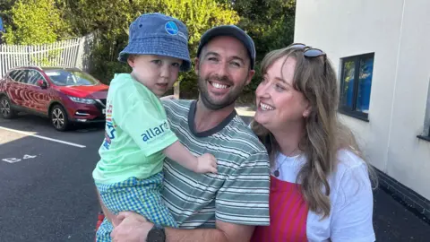 BBC News Hopcyn's dad Marc holds the young boy in his arms, with Carys standing next to them and smiling. Hopcyn wears a green t-shirt and checked trousers and a denim bucket hat, Marc wears a cap and striped shirt, and Carys wears a white shirt and red and pink striped dungarees.