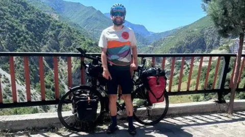 Simon Watts Mr Watts smiles for a photo against a backdrop of mountains. His bike is also in the picture and stacked with items. Mr Watts is wearing a cycling helmet and sunglasses.