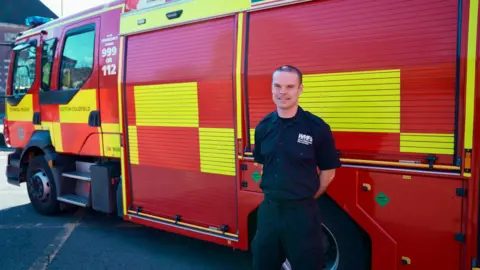 A firefighter in dark blue clothing stands beside a red fire engine with red and yellow panelling details on the side. He has a buzz cut and is stood with his hands behind his back.
