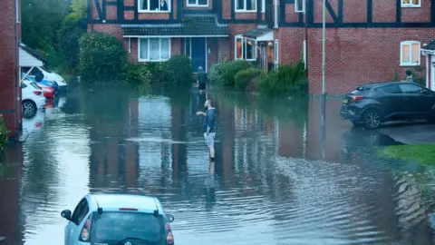 Kev Humble Photography View from a window on to a flooded road in Cheltenham. Two men are standing ankle-deep in the water, assessing the damage.