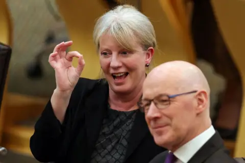 Getty Images Robison smiles and makes a circle shape with her fingers, alongside John swinney in the Holyrood chamber.