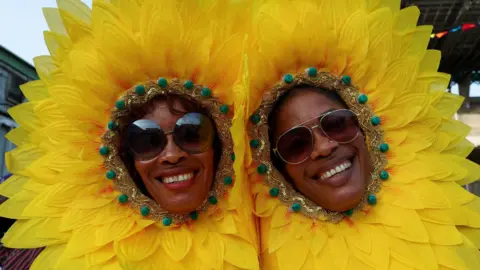 Two women, dressed as yellow flowers, smile at the camera, wearing sunglasses and petals