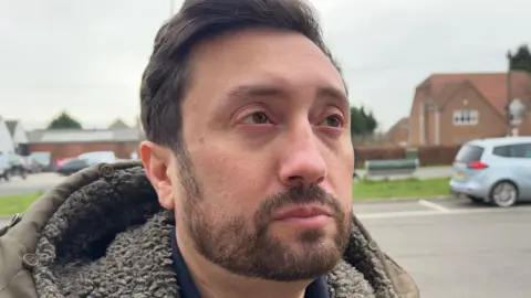 A man with brown hair, combed back, and a neatly trimmed beard, stands in a car park beside a road with residential and commercial buildings in the background. He is wearing a green parka-style coat.