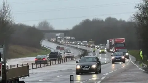 Flooding on the A30 near Ottery St Mary. There are cars and long tailbacks on the left side of the road, and cars in flood water on the opposite side.