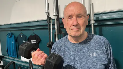 Ryan Hawthorn is 90, he has a bald head and is wearing a grey flecked t-shirt, in his right hand is a dumb bell and is bringing up towards his chest by bending his arm, behind him are various parts of lifting bars in a gym.