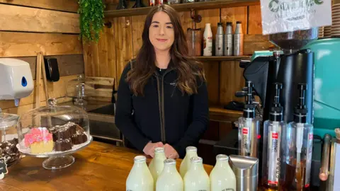 A woman with long brown hair stands behind a wooden café counter with cakes, bottles of milk, and coffee equipment. She is smiling at the camera.
