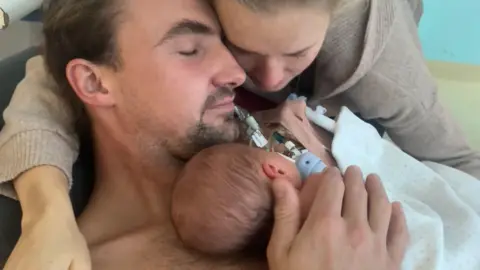 PA Media Newborn baby Poppy in hospital lying on a man's chest. A woman hugs the man. 