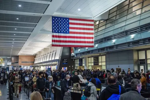 Anadolu via Getty Images Long lines of people with suitcases wrap around an airport terminal. An American flag hangs overhead. 