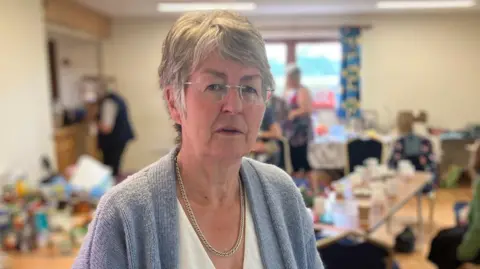 A grey-haired woman wearing a grey cardigan and white blouse, stood in a community hall with tables and chairs behind her. She is looking at the camera.