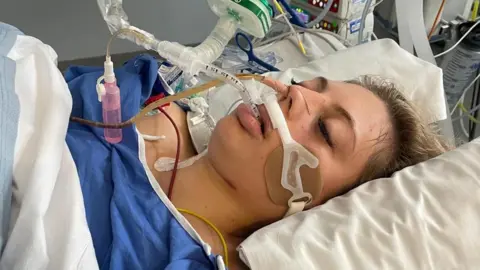 A woman lying down on a hospital bed with tubes and various medical devices connected to her. She is wearing a blue hospital gown. 
