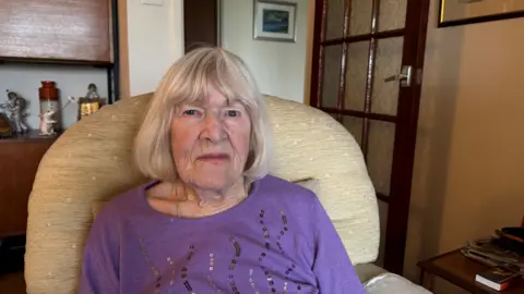 Lady with white hair, wearing a purple top and sitting on an armchair. 