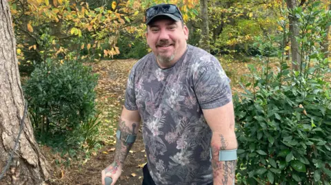 BBC Dan Stone standing in Woodland smiling at the camera. He is wearing a flowery T-shirt and has tattoos on his forearms. He also has a cap and a short goatee beard. He is using crutches because of his knee injury.